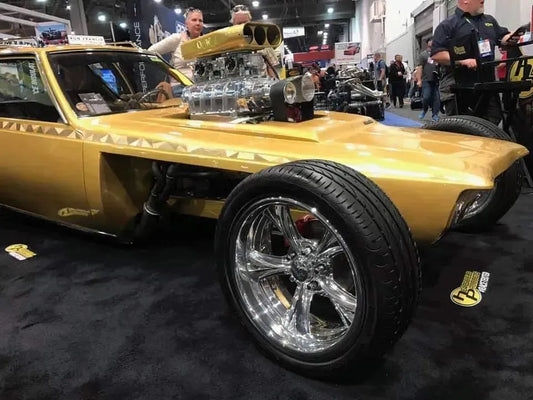 Gold custom hot rod car with large chrome wheels and exposed supercharged engine at indoor auto show