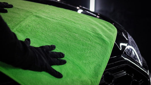 Black-gloved hand pressing bright green microfiber drying towel on glossy black car hood under studio lighting