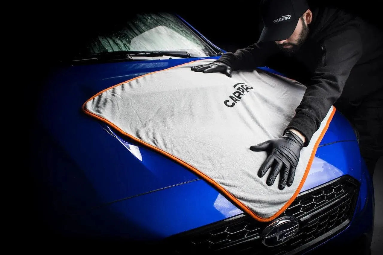 Person wearing black gloves and CarPro cap drying a blue Subaru car hood with CarPro microfiber towel