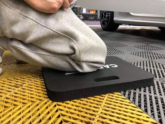 Person kneeling on a black CarPro foam kneeling pad in a garage with a white car nearby