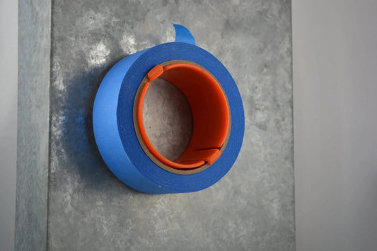 Blue painter's tape roll with orange dispenser on textured metal surface