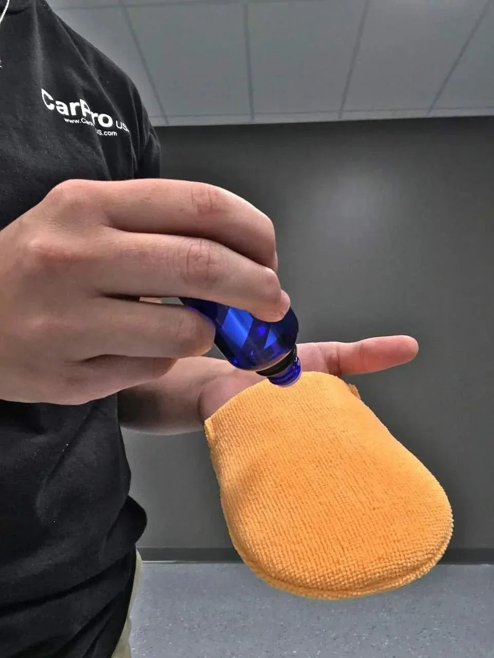Hand holding orange microfiber applicator mitt while applying liquid from blue bottle, indoor setting