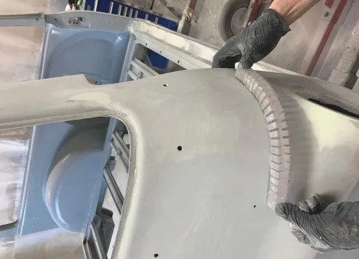 Hands wearing gloves holding a curved linear blocking tool over a gray primer-coated car body in a workshop